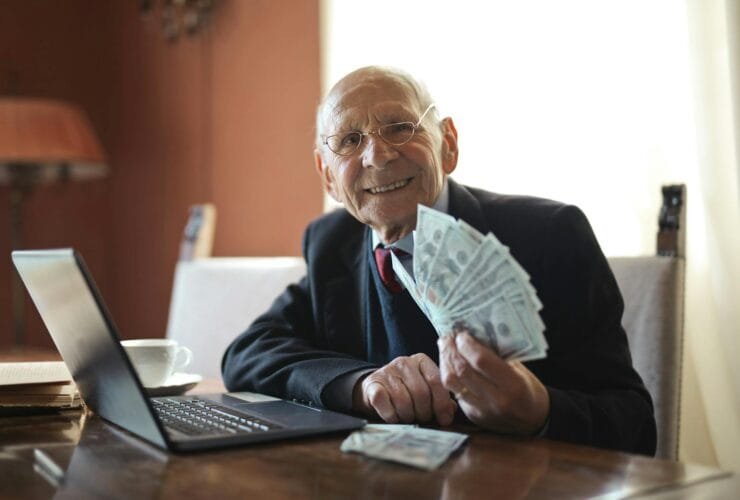 happy senior businessman holding money in hand while working on laptop at table